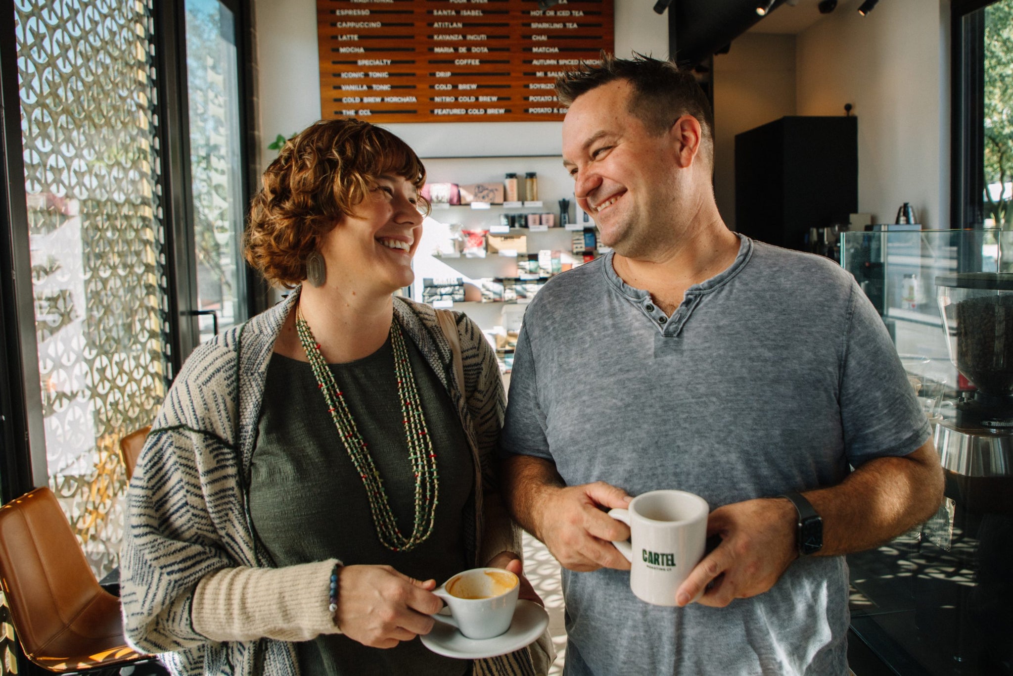 Smiling woman and man hold coffee cups inside a Cartel Roasting Co cafe