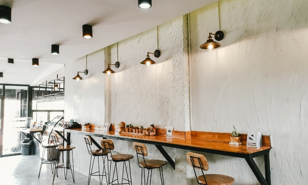 Bright modern coffee shop interior with a long wooden counter, bar stools, and a white textured wall.