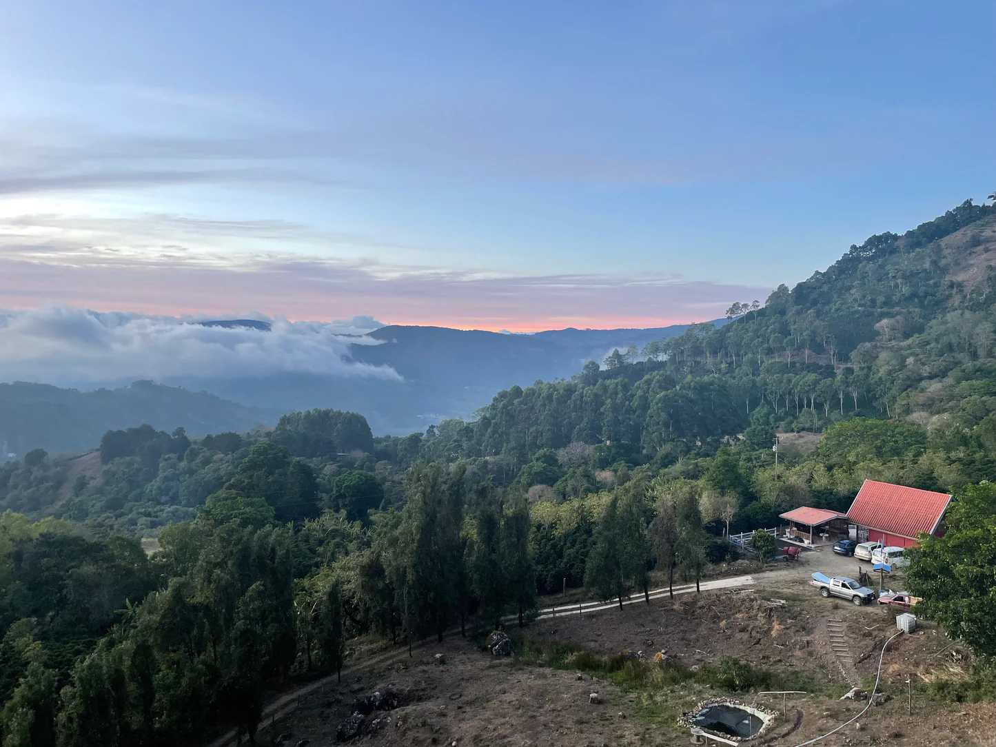 Panoramic view of a forested valley at sunset with low clouds and a red roofed building on the hillside.