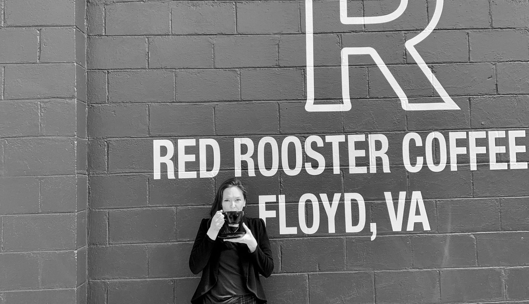 Person holding a coffee cup in front of a mural reading Red Rooster Coffee