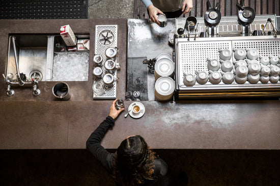 Overhead view of a cafe counter with a barista pouring milk beside an industrial espresso machine