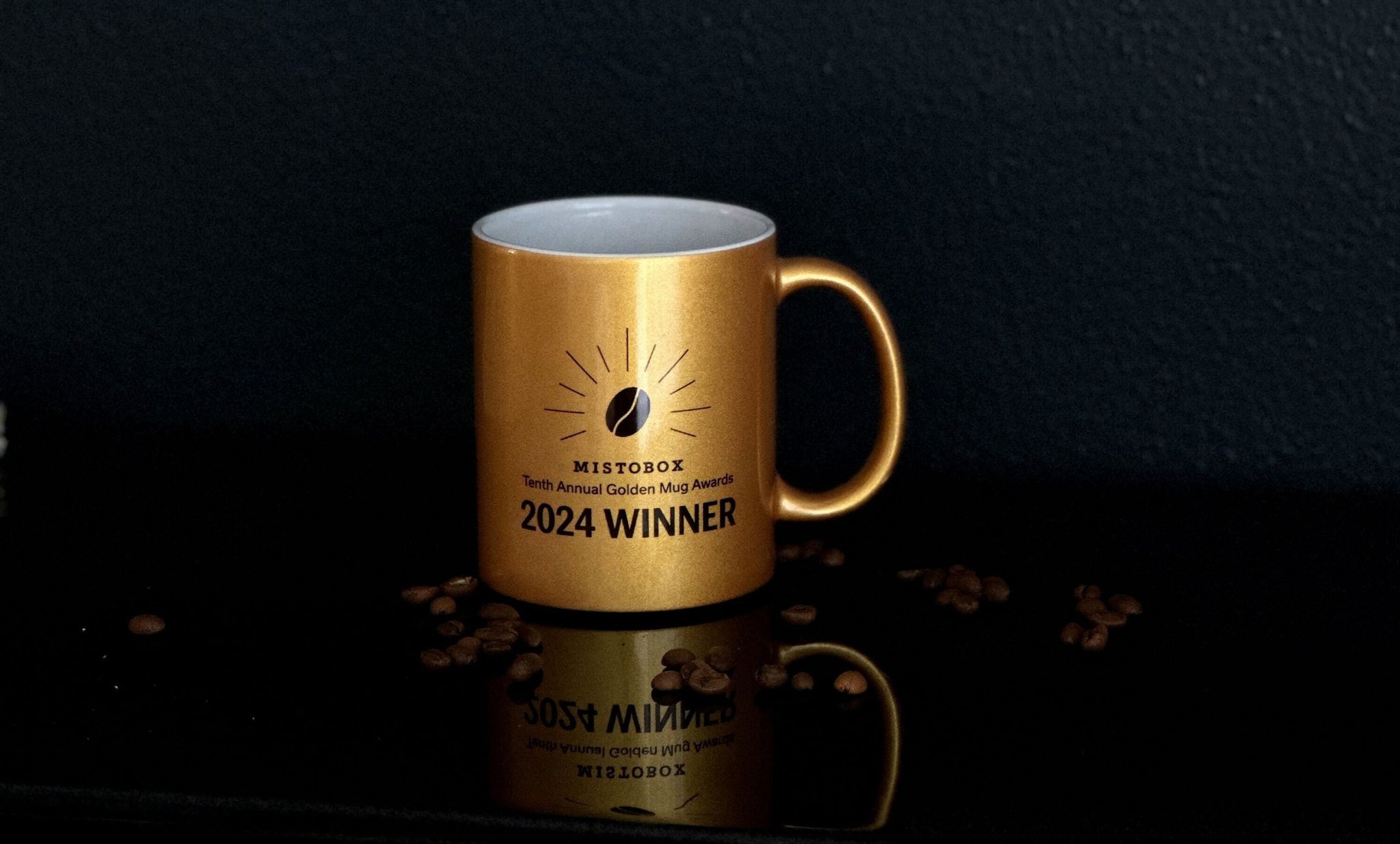 Golden mug displaying 2024 WINNER and Mistobox branding on a dark reflective surface with coffee beans around
