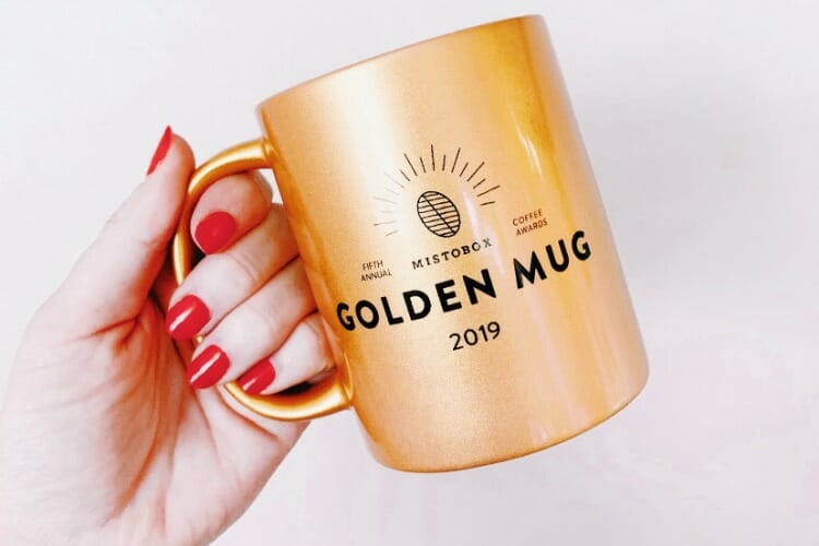 Shiny gold mug labeled Golden Mug 2019 held by a hand with red nails.