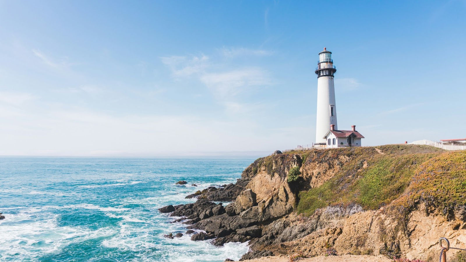 Coastal scene with a white lighthouse atop a rocky cliff overlooking the blue ocean under a clear sky