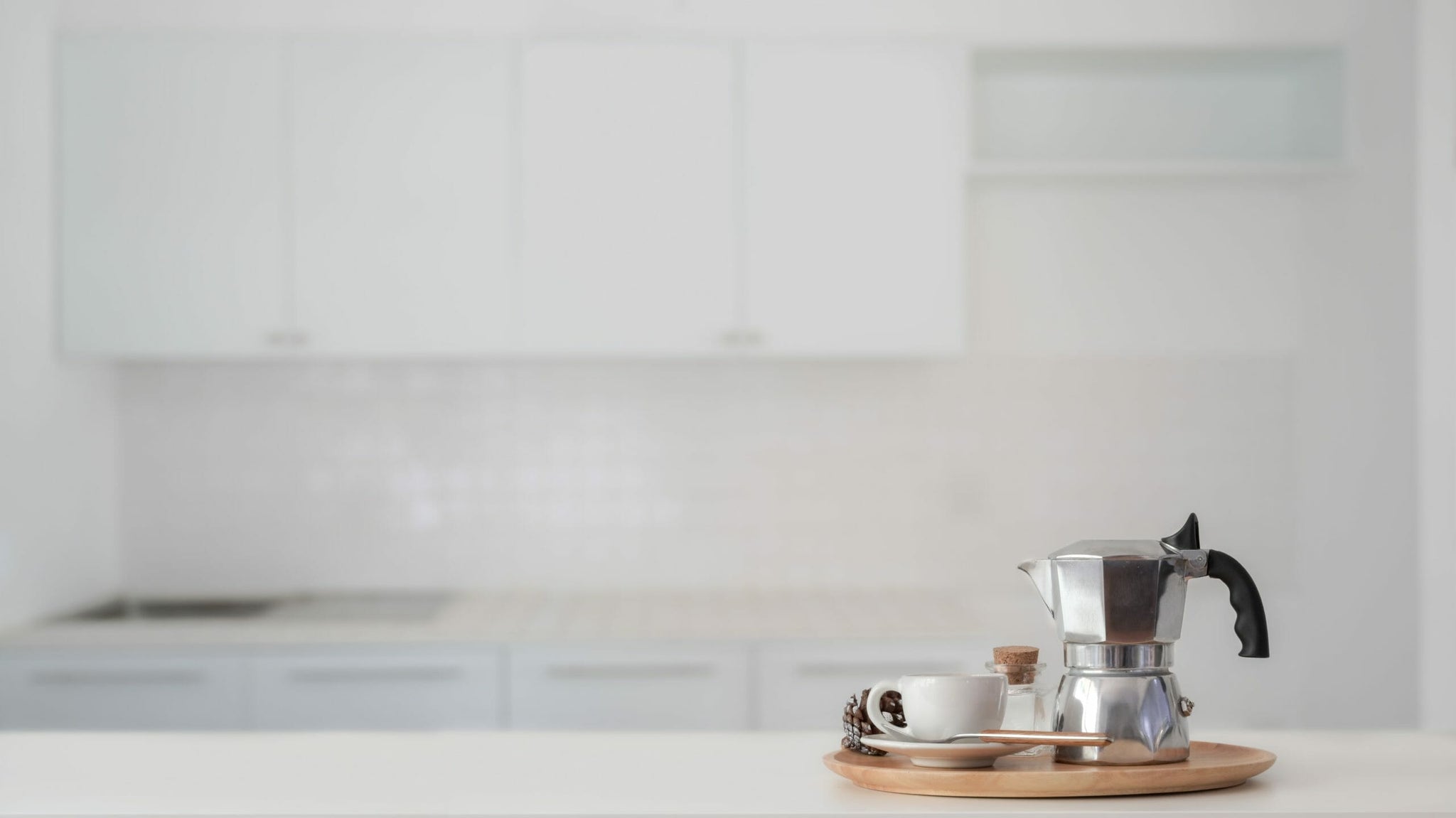 Stovetop moka pot on a wooden tray with cups in a bright white kitchen.