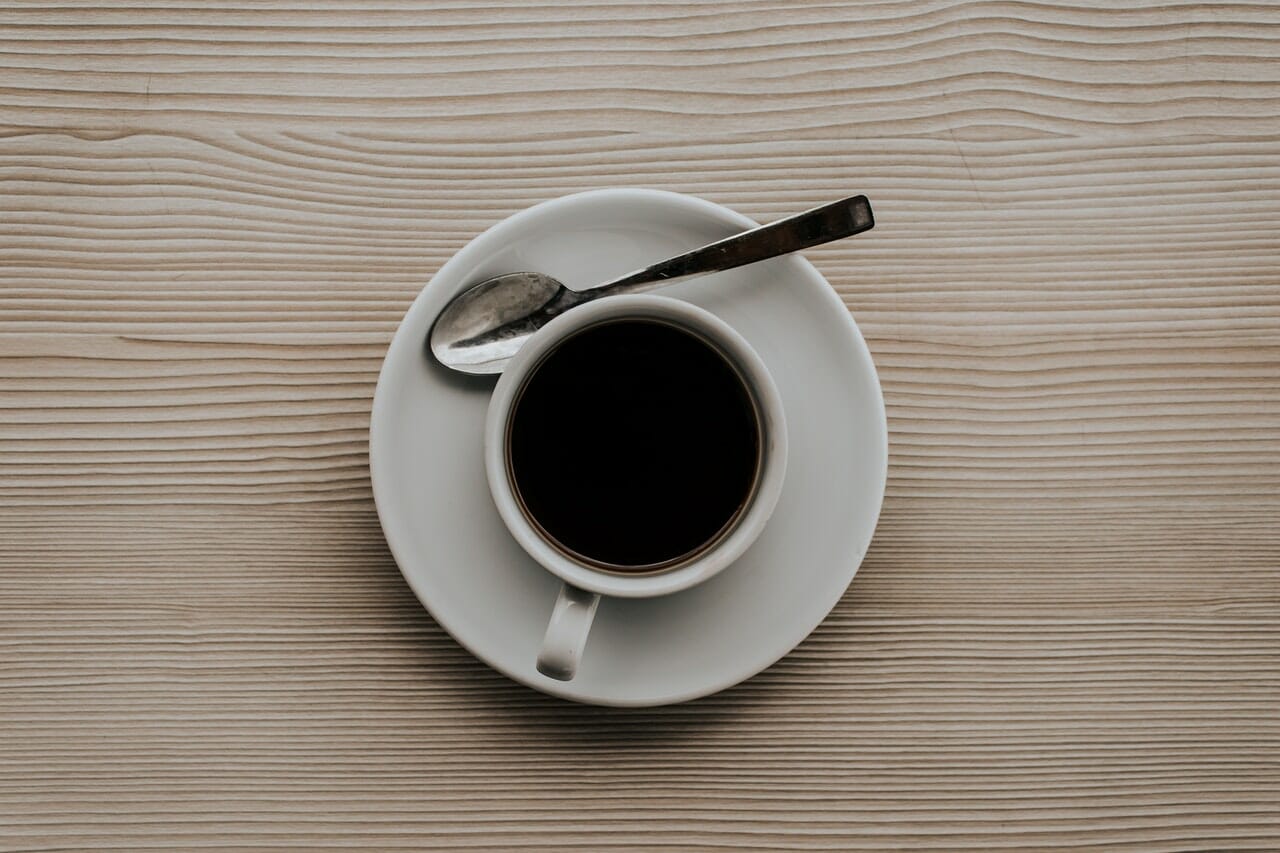 Top-down view of a white cup of black coffee on a saucer with a spoon beside it on a light wooden table.