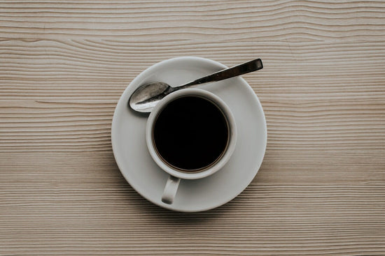 Top-down view of a white cup of black coffee on a saucer with a spoon beside it on a light wooden table.