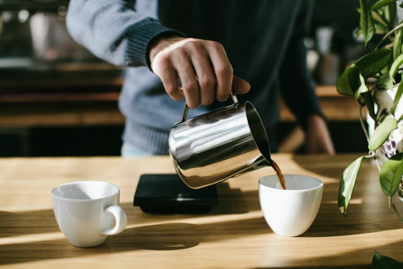 A hand pours coffee from a metal kettle into a white cup on a wooden table.